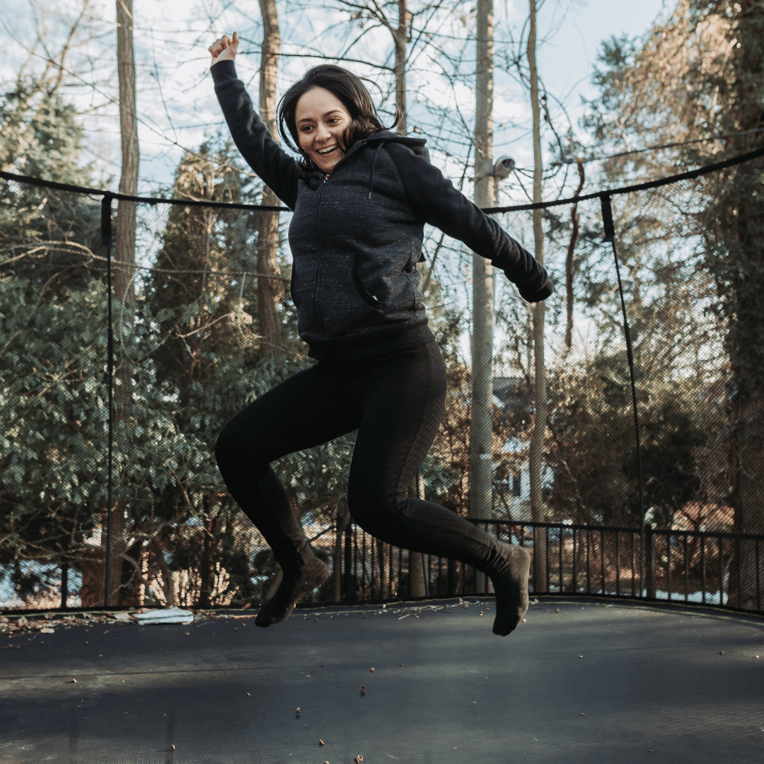 leadership-and-resilience-bounce-back A woman bouncing on a large trampoline in the garden. She is wearing a jacket but no shoes. Her hands are in the air and she looks triumphant.