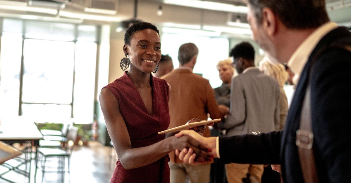 A woman in professional clothing smiles and shakes hands with a man out of shot. She is at a stylish networking event.